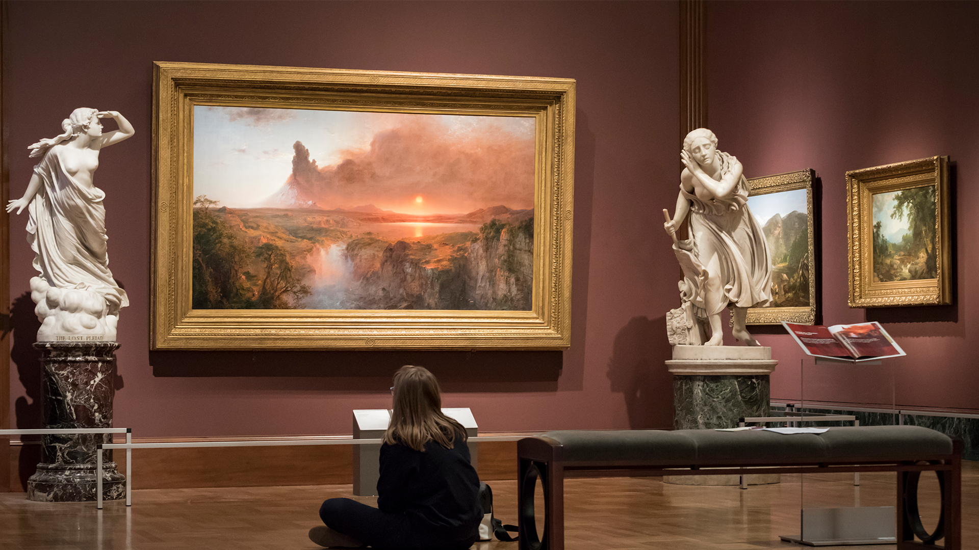 A visitor sitting on the floor next to a bench in front of Frederick Church's "Cotopaxi"